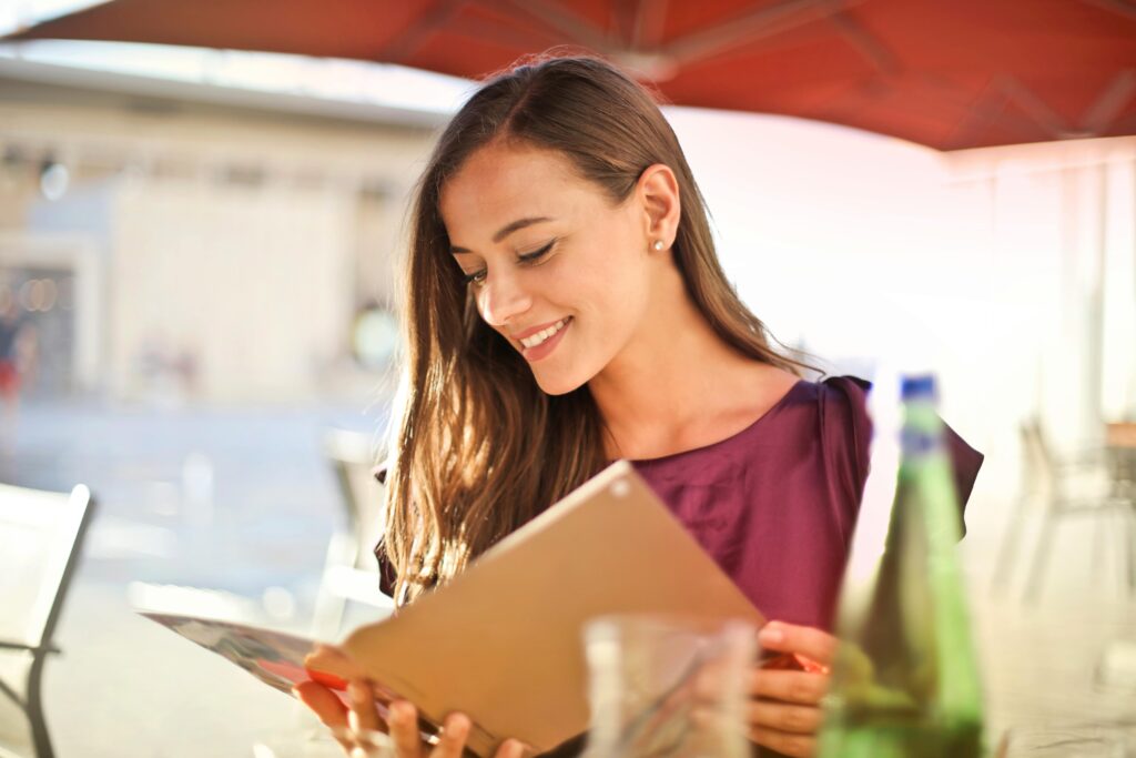 A woman smiles as she reads a menu at an outdoor restaurant, enjoying a sunny day.